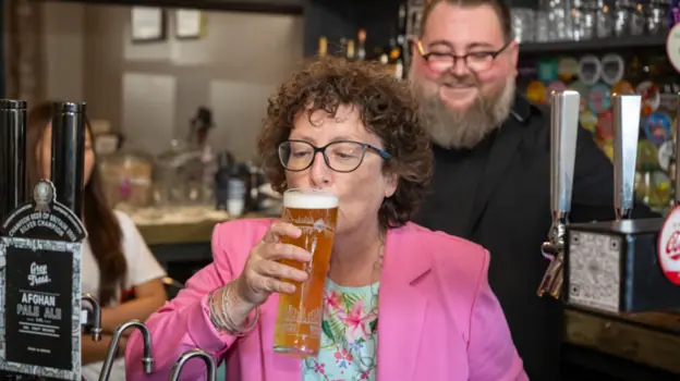 Wearing a pink blazer, the Welsh government's finance minister Elin Jones sips a freshly-poured pint behind the bar at the Radyr Tap. There is a man with a beard and glasses, with a black shirt stood…