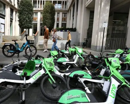 Two women and a man pass in single file in front of a Forest ebike on the pavement and about 10 Lime ebikes lying on top of each other in the street