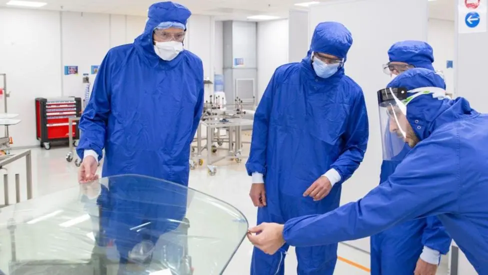 Four workers in blue overalls inspect an aircraft window. They are wearing masks and hoods.