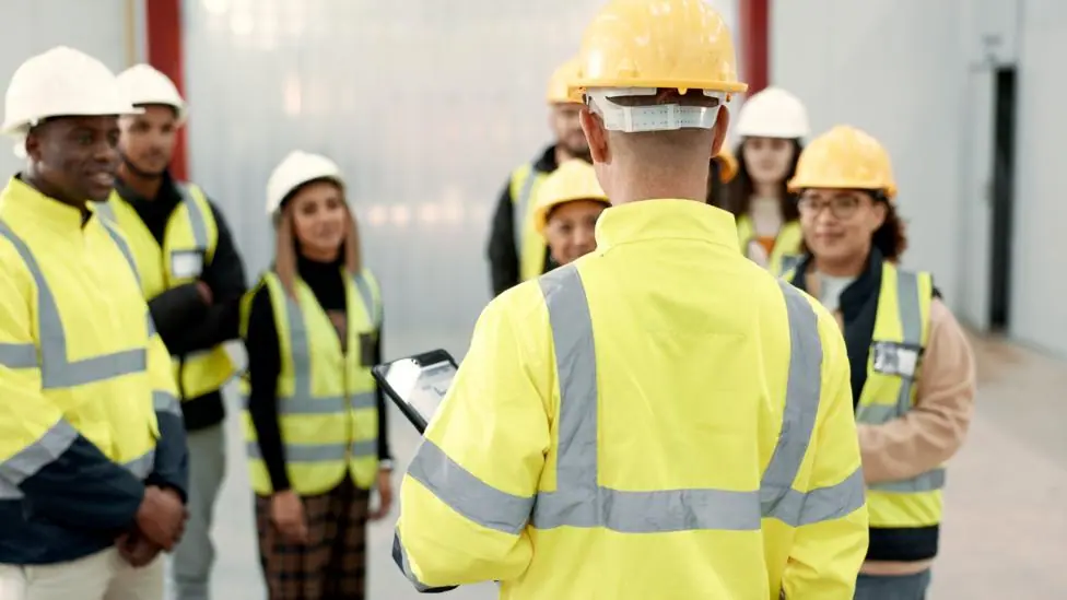 A group of people wearing hi-vis yellow jackets stand around in a room. Most of the group are standing facing a man with a clipboard, whose back is towards the camera. They are listening to what he…