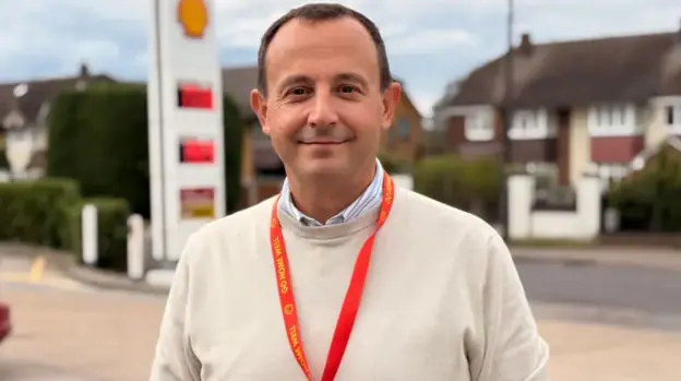 Man looks at camera, wearing beige jumper over a shirt
