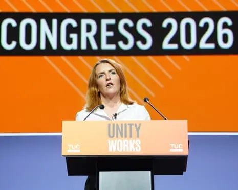 A woman stands behind a podium, with the words 'unity works' on it, speaking into a microphone.