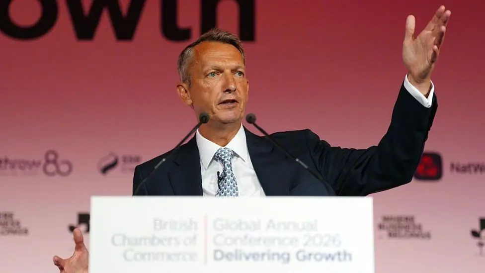 Andy Haldane gestures animatically as he speaks from a white podium in front of a red background at a British Chambers of Commerce event.