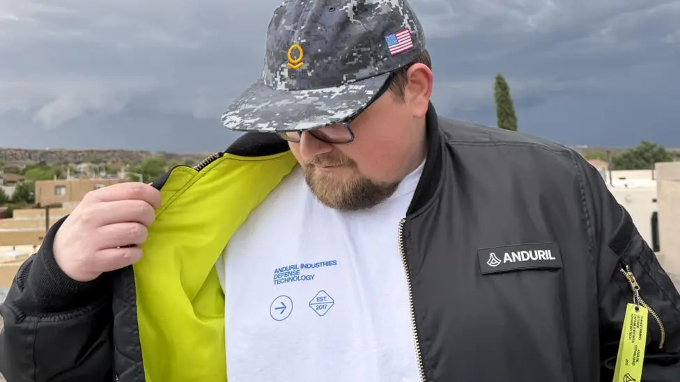 Jonathan Sargent wearing a t-shirt and jacket from US defence firm Anduril and a hat from US software firm Palantir