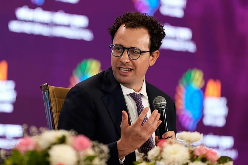 Anthropic boss Dario Amodei sitting and speaking at a conference, holding a microphone and wearing a suit with a purple tie