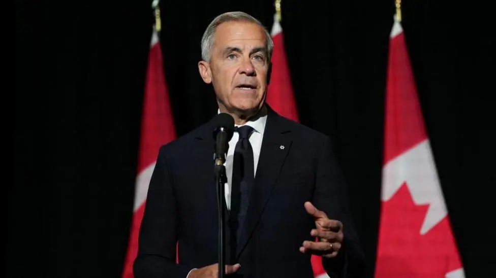 Mark Carney, in a black suit and tie gestures as he stands in front of a microphone. Behind him, against a dark background, are three red and white Canadian flags in a row.