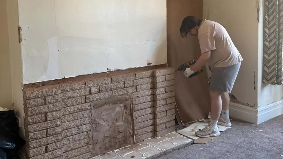 A man in shorts and a t-shirt, using a power tool to refurbish a fireplace.