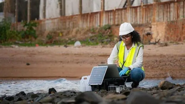 A woman in a yellow hi-vis jacket, a white hard hat, white shirt and jeans crouches on some rocks next to a stream of water next to a red sandy bank. In the background is a factory behind a high fence
