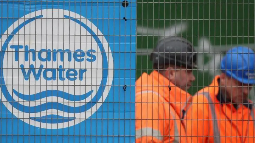 Two men in orange hi-vis jackets and safety helmets stand behind a wire fence. To their left is a blue and white Thames Water logo.