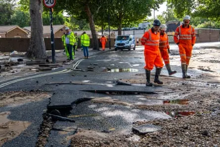 Men in orange hi-vis overalls and wellies stand by puddles in a road where the surface is cracked and potholed. Others in yellow vests stand in the background.