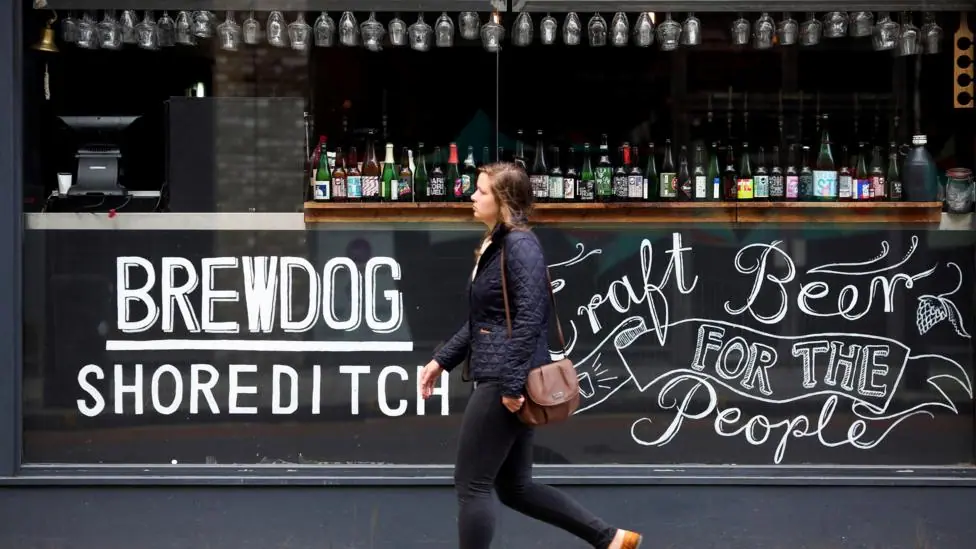 A woman walks past a Brewdog bar in London. On the glass is written Brewdog Shoreditch and Craft Beer for the People.