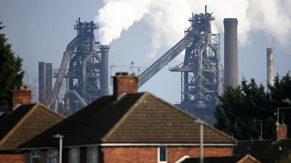 A British Steel plant, with steam billowing out of it, behind a row of terrace houses