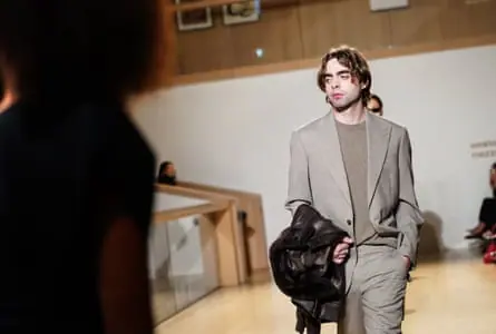Lennon Gallagher on the catwalk wearing a grey suit and holding a jacket over his arm