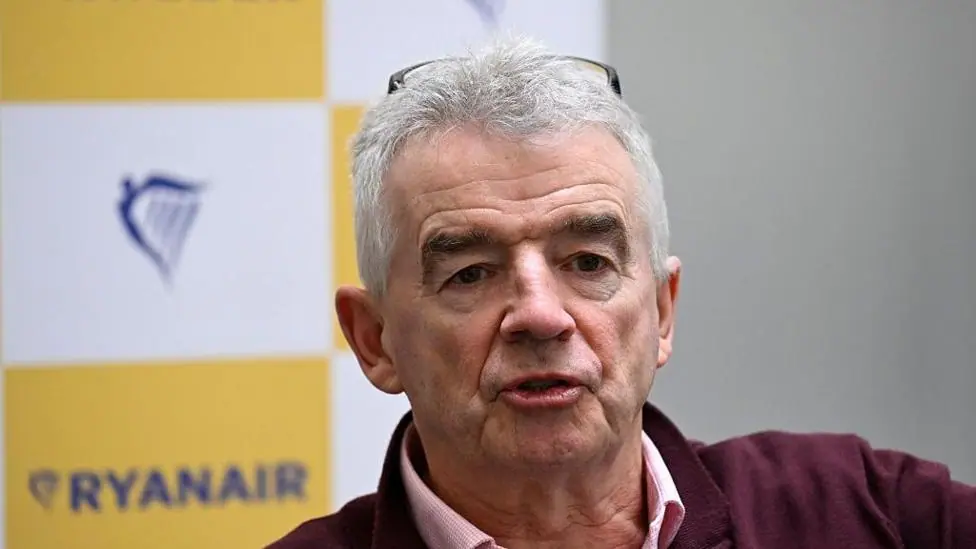 A close-up Michael O'Leary, chief executive of Ryanair, wearing a pink and white chequered shirt and burgundy jumper. Behind him, the Ryanair logo is on a printed background in soft focus.