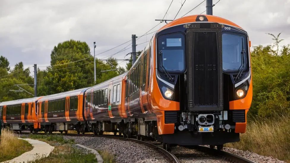 A dark orange train travels across train tracks in the countryside with trees on its left side.
