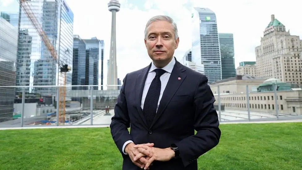 Champagne wears a suit and tie while he stands on a lawn in front of the Toronto city skyline of high-rise buildings.