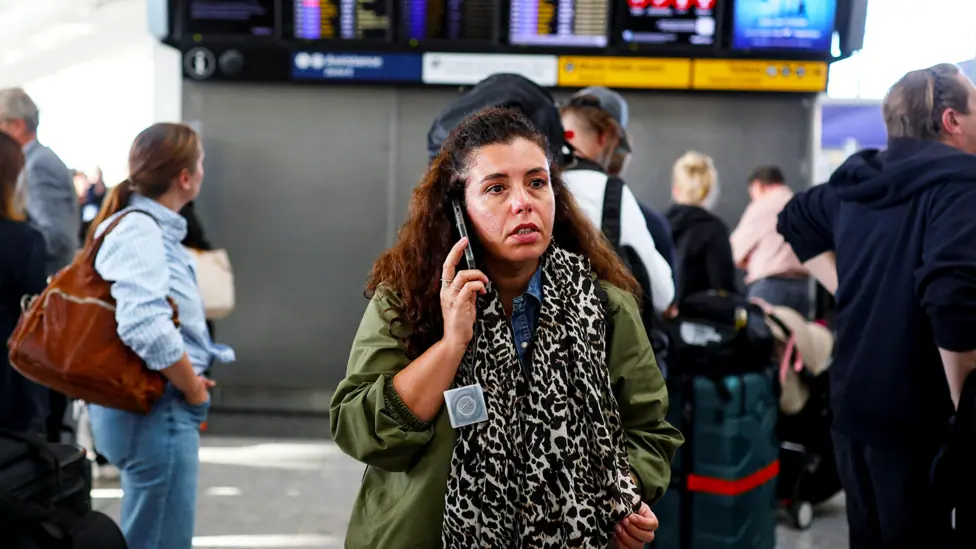 A woman on the phone at Heathrow with a departure board in the background showing delays and cancellations due to air traffic control problems