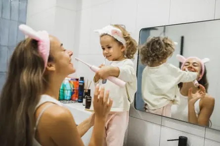 A toddler stands up at the sink and brushes her mum’s teeth with an electric toothbrush. Both are wearing fluffy bunny ears