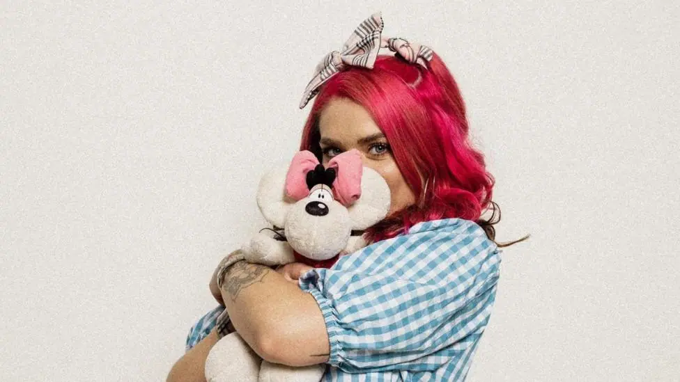 Woman in blue and white checked shirt with dyed pink hair and a tartan hair bow hugging white teddy.