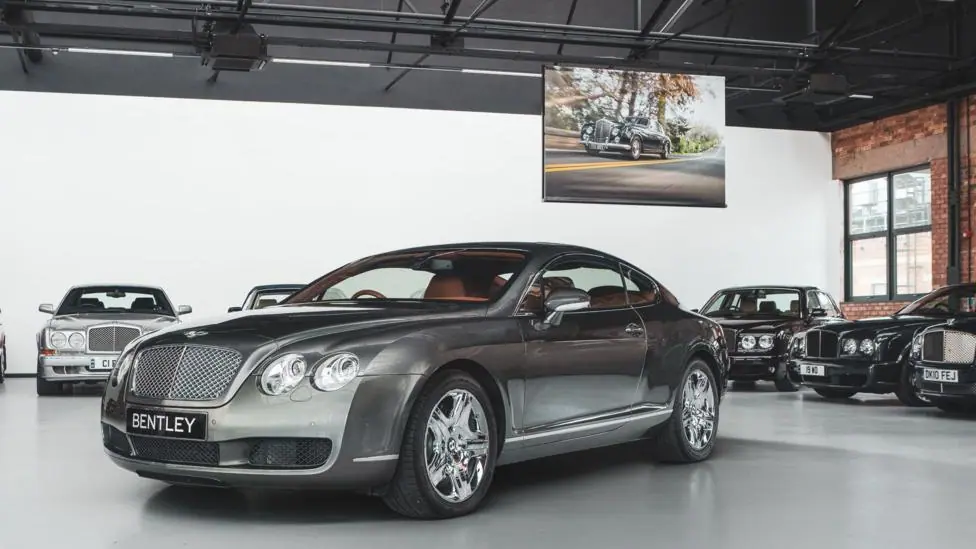 A Bentley showroom with a saloon car in the foreground and other cars lined up against the walls around it. A hanging screen with an image of an older car on a road is hanging from the ceiling behind…