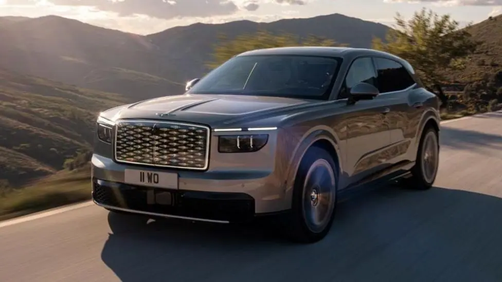 An aluminium-coloured Bentley SUV is driven on a road with hills and countryside behind it.