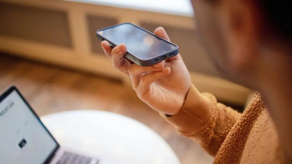 Stock photo shows a person speaking into a phone while sitting at a desk indoors on their laptop.