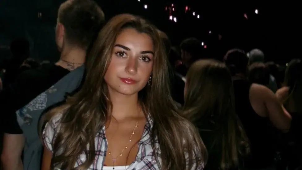 Evie Lewis, a young woman standing in what appears to be a music venue. She has long dark hair and is wearing a checked short sleeved top over a white vest with a thin gold necklace. Behind her there…