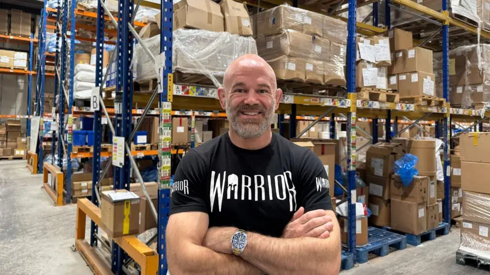 Kieran Fisher, a bald man with a short grey beard wearing a black T-shirt with the word WARRIOR on the front, standing smiling in a warehouse. Behind him are cardboard boxes stacked on blue metal…