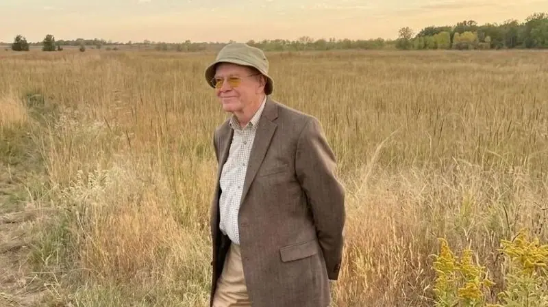 Lawyer BF Hicks stands in the middle of a field. He is wearing a wool formal jacket and a hat