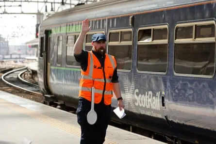 A train guard in an orange hi-vis vest blows his whistle for departure as a Scotrail train departs from Glasgow Central Station.