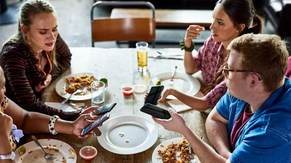 Three young people viewed from above are sitting at a table at the end of a meal. They have their phones out and the woman on the left is frowning as she looks at hers. There are empty and half empty…