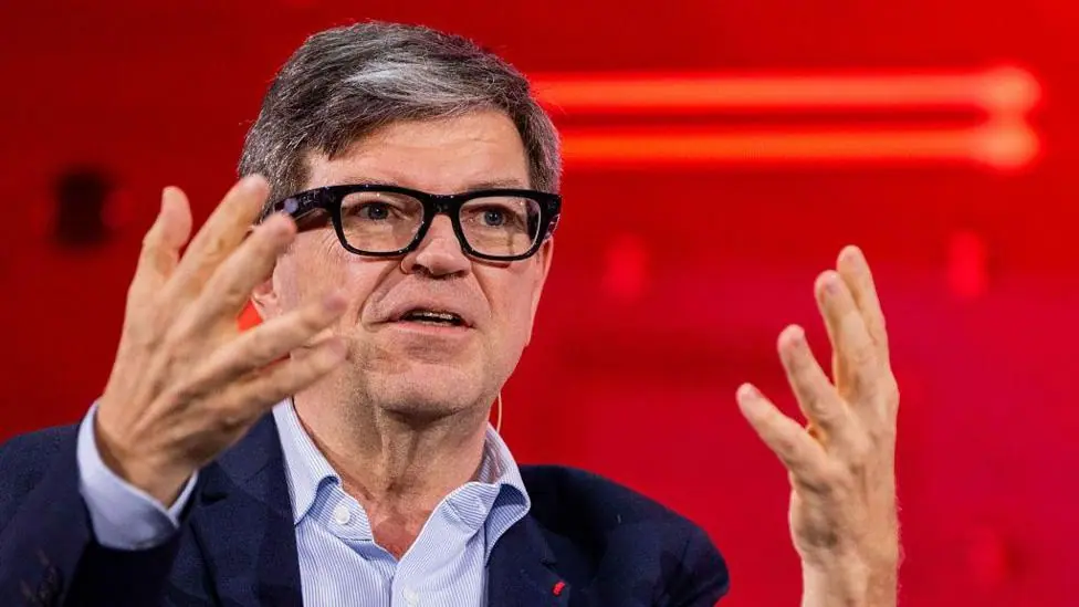 Yann LeCun wears fashionable thick-rimmed glasses, a light blue shirt and navy jacket. He is using his hands to articulate a point while speaking at a conference.