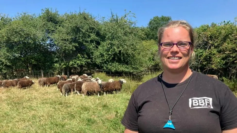 Abby Cook, a woman with blonde hair, glasses and a dark top, is standing in a grassy field in front of a flock of sheep with dark wool.