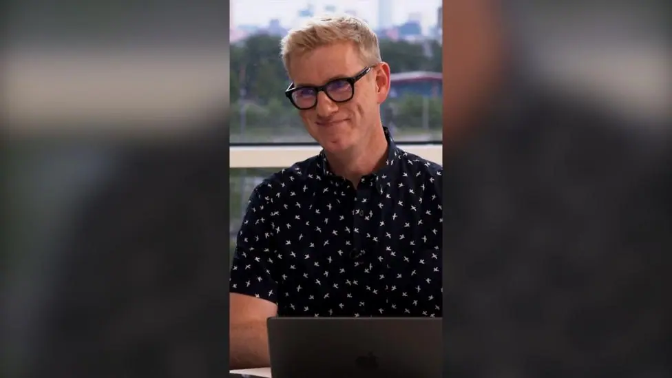 A man with glasses and blonde hair smiles and looks to the side of the camera. He has a laptop in front of him.