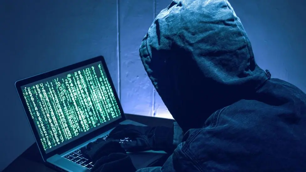 A man wearing a black hoodie and black gloves has his back to the camera. His hood is up and you cannot see his face. His in a dark room and is working on a laptop which has lines of green numbers…