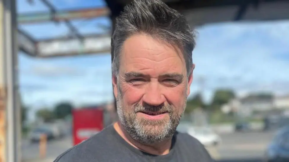 Sean is looking at the camera and is wearing a grey t-shirt. He has black hair and a greyish beard.