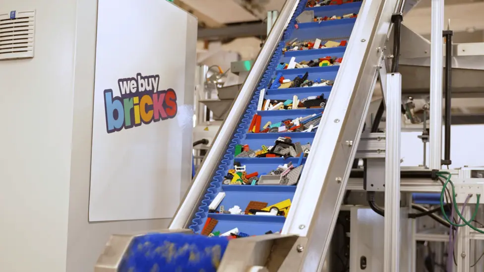 Multicoloured Lego pieces travel through a blue conveyor belt beside machinery bearing the WeBuyBricks logo.