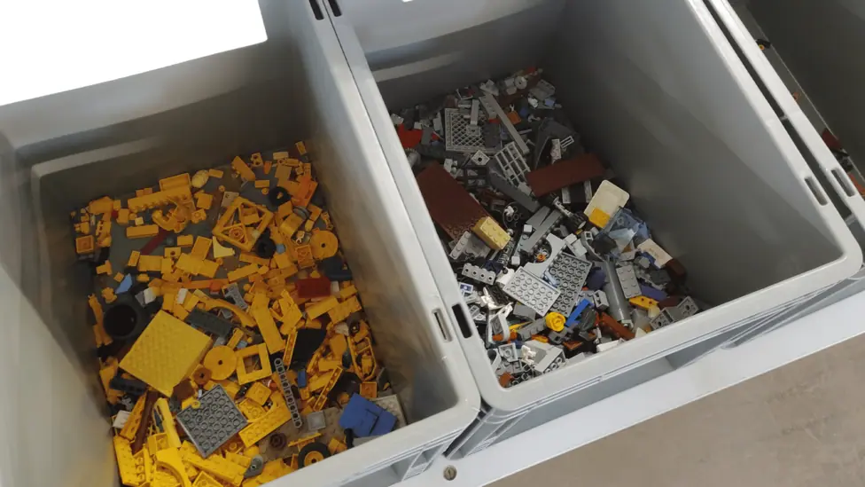 Large grey containers hold separated groups of Lego, including yellow pieces and mixed grey and coloured pieces.