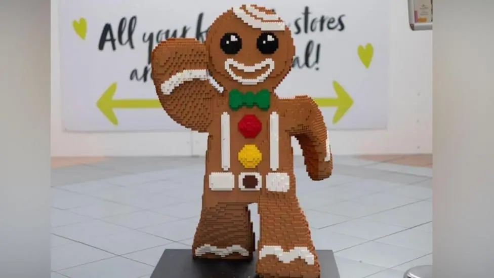 A large figure of a gingerbread man created out of Lego bricks. The colouring is brown with round black eyes, and a white outline of a mouth, and one arm raised. A green bow tie, and a red and yellow…