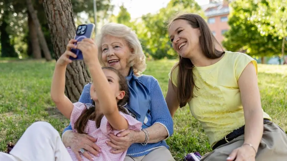 A young girl takes a selfie on a mobile phone as she lies on the lap of a woman who looks like her grandmother, and another woman who represents the girl's mother smiles next to them.