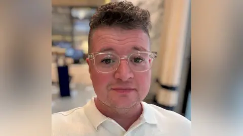 A person with a short brunette quiff and thick dark eyelashes wears a white shirt and clear framed glasses in a selfie.