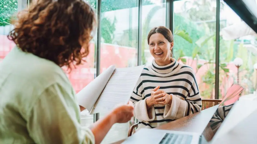 Two women are talking. One is holding a piece of paper while the other is talking.