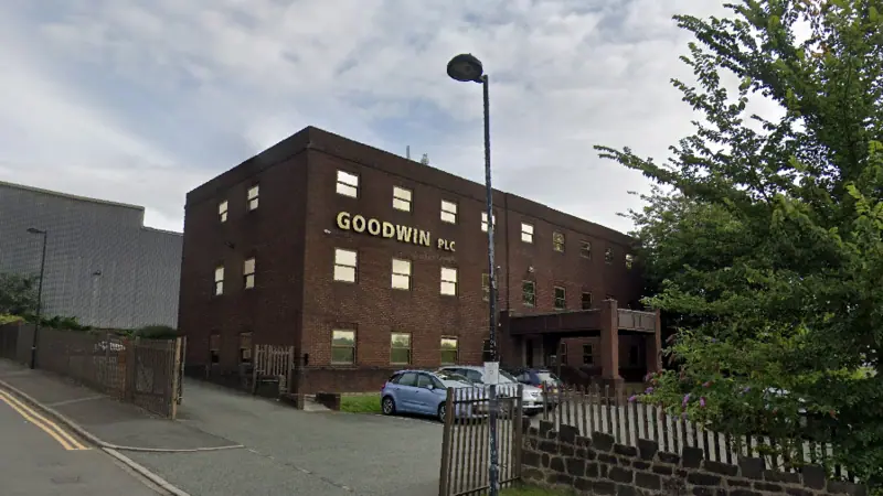General view of a three-storey brown-brick rectangular building with three cars parked at the front. On the right of the image is a tree next to a wall