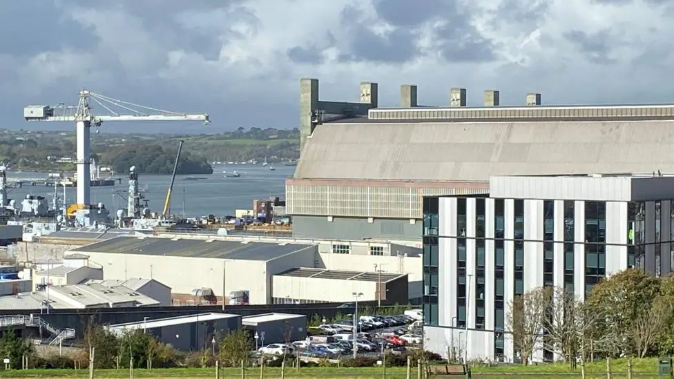 A general view of of the dockyard. Large buildings and a crane are visible. Water and boats are in the distance. The sky is grey and cloudy.