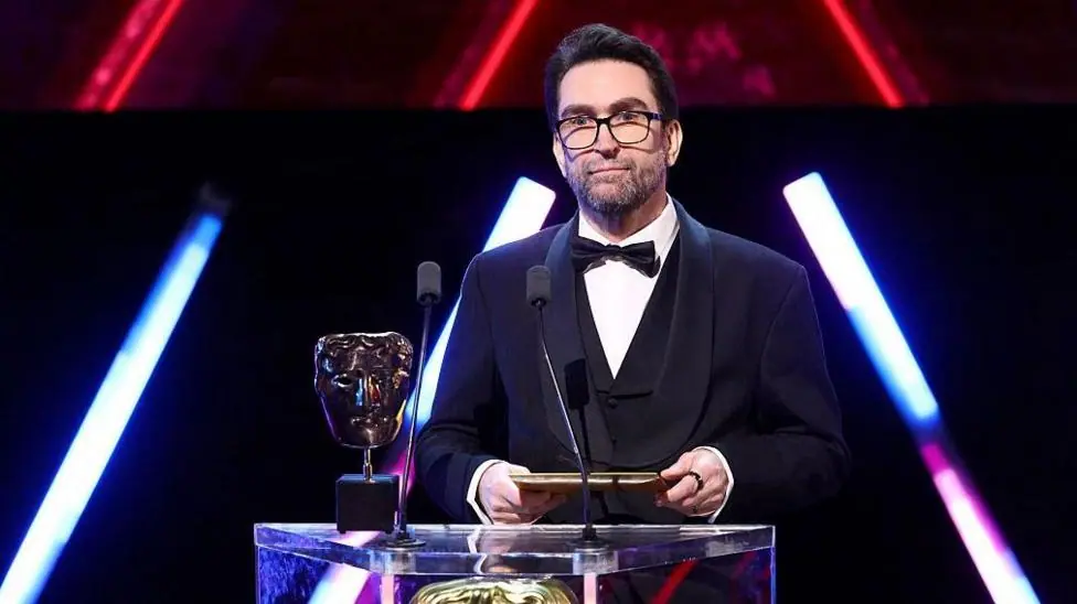 A man wearing glasses and a three-piece suit with bow tie stands on-stage behind a lectern, holding an envelope in both hands. On the lectern are two microphones and a Bafta award - a golden trophy…