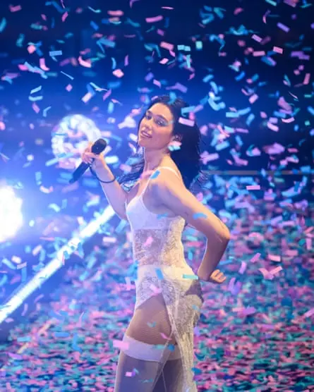 Dua Lipa holds a microphone and wears a lacy white dress with confetti falling around her