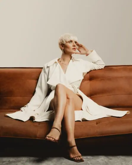 Millar in a white trench coat seated on a brown sofa