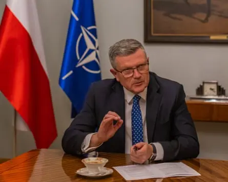 Marcin Bosacki pictured in a suit behind a wooden table with the Polish and Nato flags in the background