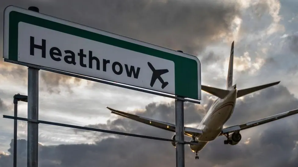 A commercial aeroplane flying low on final approach to land at Heathrow Airport, passing directly over an overhead highway road sign.