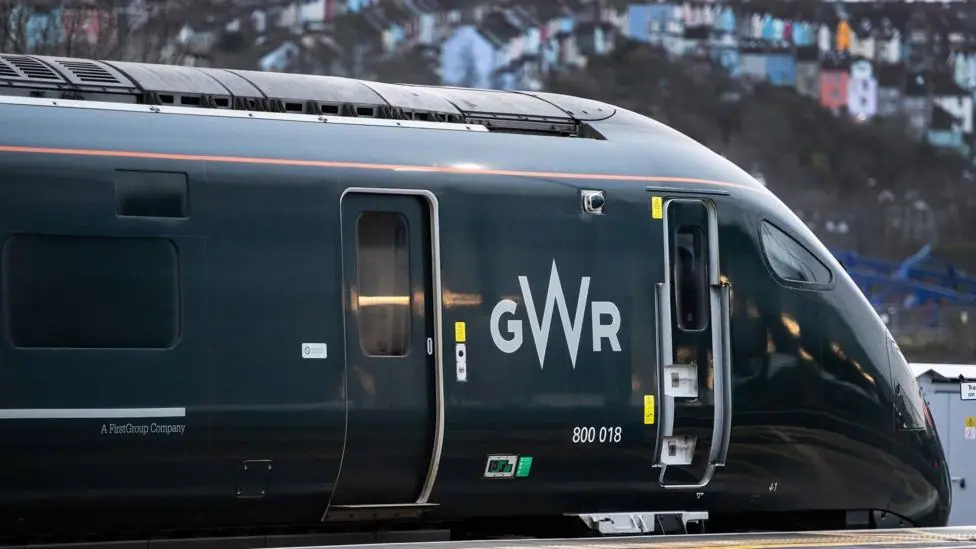 A green train at a platform, with colourful houses slightly out of focus in the background.